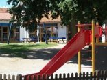 Außengelände eines Kindergartens. Im Vordergrund wird eine rote Rutsche abgebildet und im Hintergrund ein dunkelblaue Schaukel