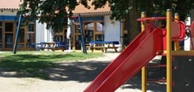 Außengelände eines Kindergartens. Im Vordergrund wird eine rote Rutsche abgebildet und im Hintergrund ein dunkelblaue Schaukel