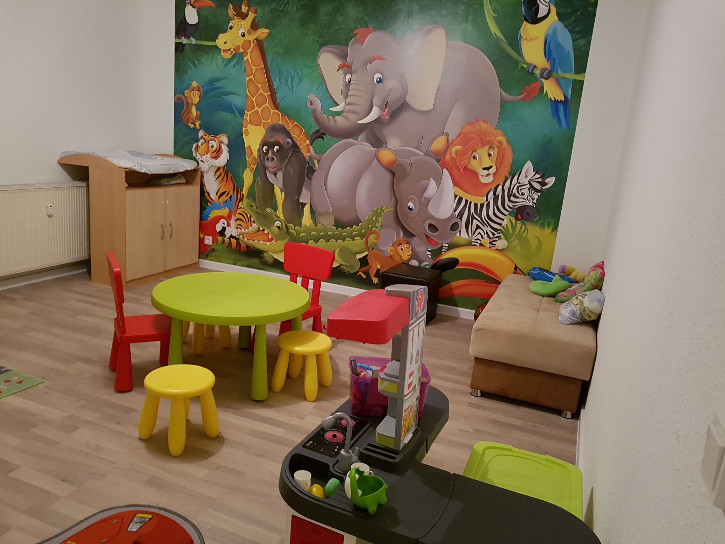 Ein kindergerechtes Zimmer mit kleinen Stühlen und einem kleinen Tisch zum Sitzen, einer Kinderspielküche und einer Kommode. Die Wand ist mit einer Fototapete beklebt, auf der einige Zootiere abgebildet sind. 