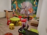 Ein kindergerechtes Zimmer mit kleinen Stühlen und einem kleinen Tisch zum Sitzen, einer Kinderspielküche und einer Kommode. Die Wand ist mit einer Fototapete beklebt, auf der einige Zootiere abgebildet sind. 