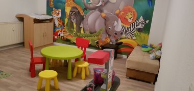 Ein kindergerechtes Zimmer mit kleinen Stühlen und einem kleinen Tisch zum Sitzen, einer Kinderspielküche und einer Kommode. Die Wand ist mit einer Fototapete beklebt, auf der einige Zootiere abgebildet sind. 