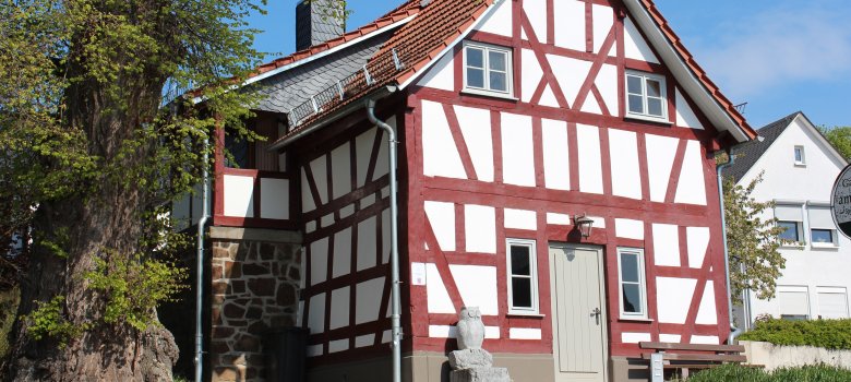 Auf dem Bild ist das Backhaus von Laufdorf zu sehen. Es ist ein altes Fachwerkhaus. Die Balken sind rot gestrichen. Vor dem Haus steht eine Eule aus Stein und links daneben steht ein großer Baum