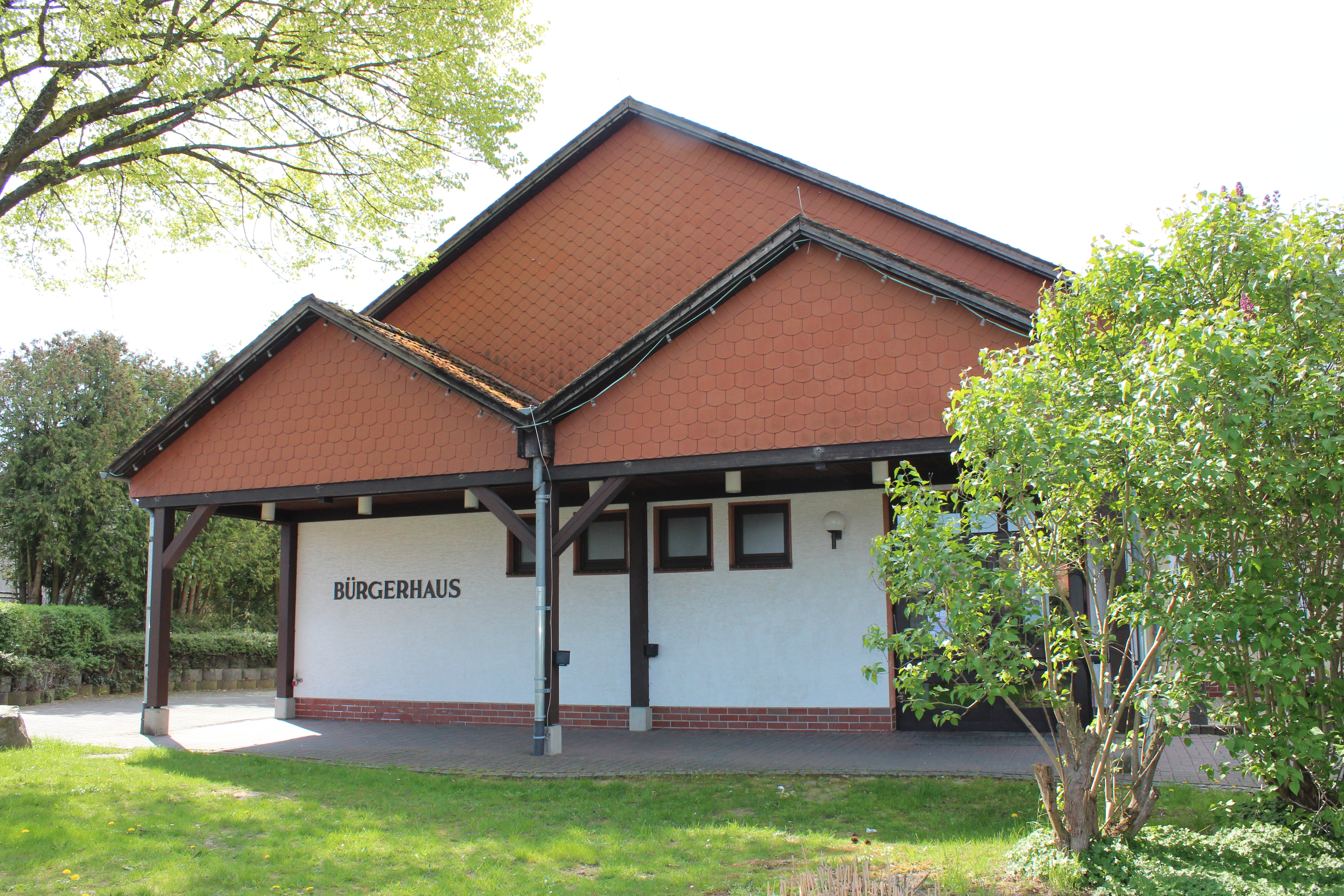 Auf dem Bild sieht man das Bürgerhaus in Schwalbach. Auf der rechten Seite ist ein Fliederbaum zusehen