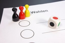 Ein weißer Zettel mit der Aufschrift Wahlen und zwei neutrale Felder zum Ankreuzen. Auf dem Zettel stehen drei Spielfiguren in den Farben Schwarz, Rot, Geld und ein Würfel deren Seiten in den selben Farben sind