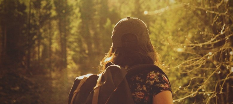 Ein Mädchen steht mit Rücken zur Kamera. Auf dem Rücken trägt sie ein Rucksack und auf dem Kopf eine Mütze. Im Hintergrund sieht man einen Wald der mit Sonnenlicht angeleuchtet wird. 